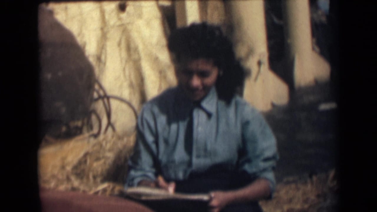 1946 India home-movie frame of a woman in a blue shirt looking down as she writes or sketches in a notebook beside straw and wooden posts.