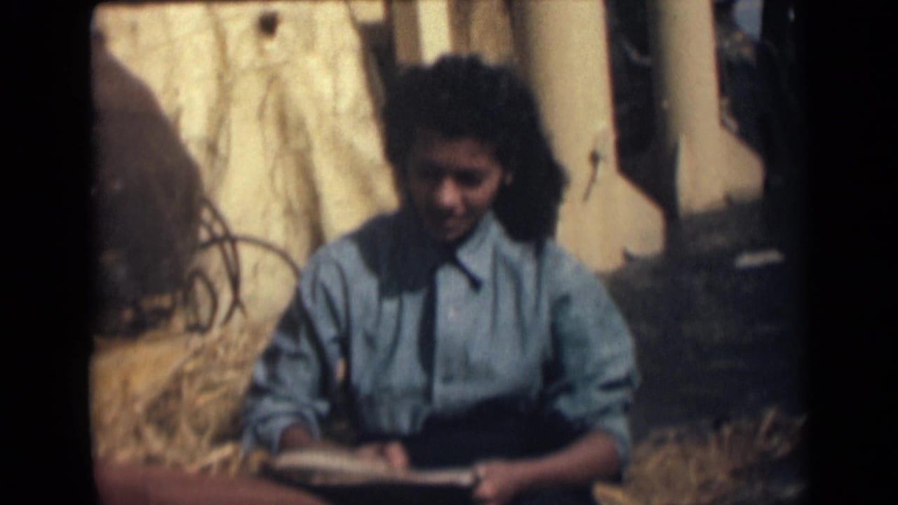 Girl seated outdoors writing in an open notebook beside dry grass and posts.