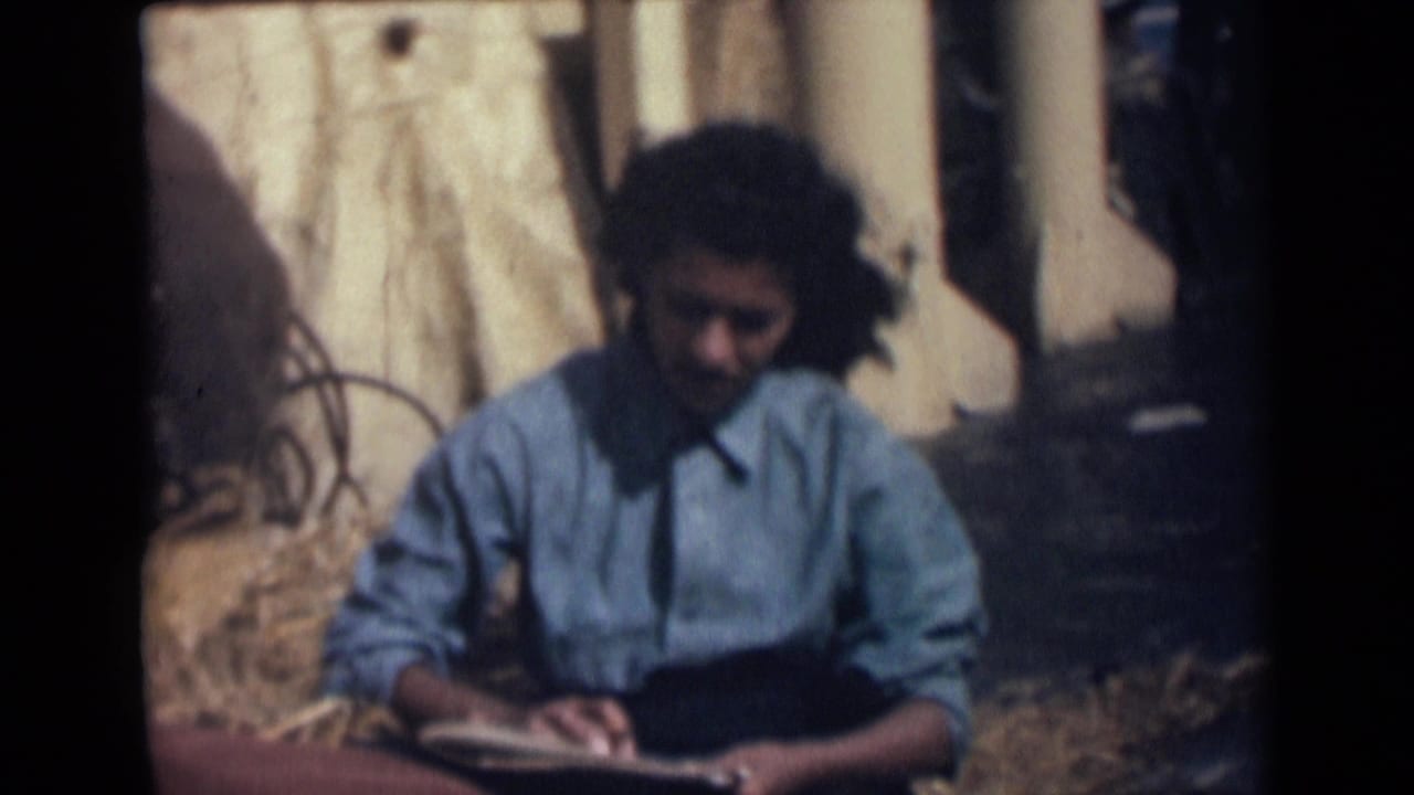The 8mm frame shows a girl seated outdoors in India, leaning toward her notebook with dry grass and structural posts behind her.