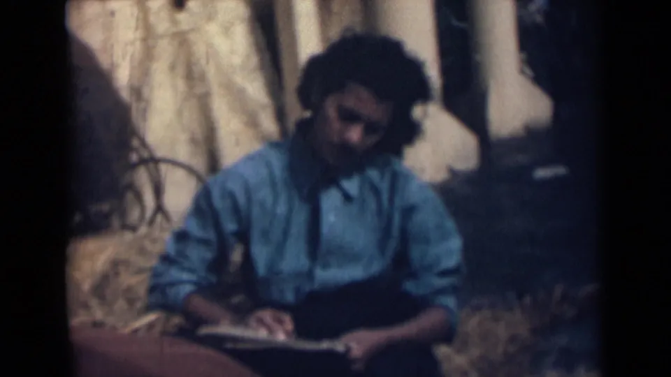 A girl in a blue shirt sits with an open notebook in 1946 India footage, with windblown hair and a rustic outdoor backdrop.