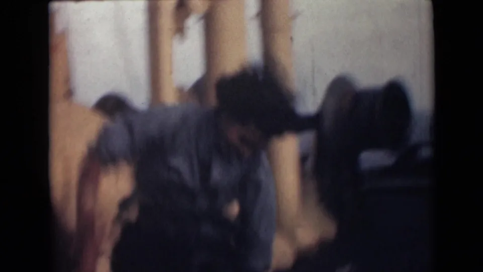The woman bends forward in soft-focus 8mm footage on a ship deck with columns and railings behind her.