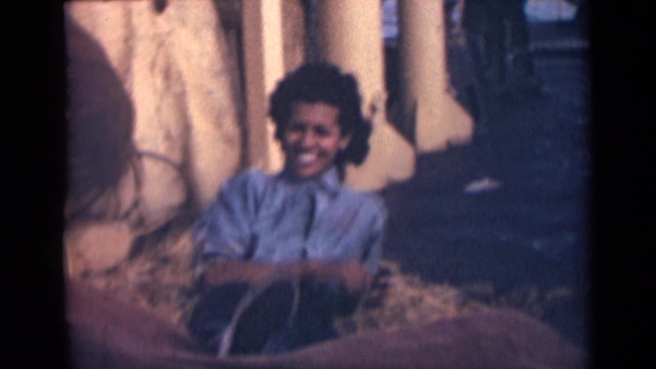 A smiling woman sits on or near the deck surface in 1946 India footage, with straw or rope and ship structures nearby.