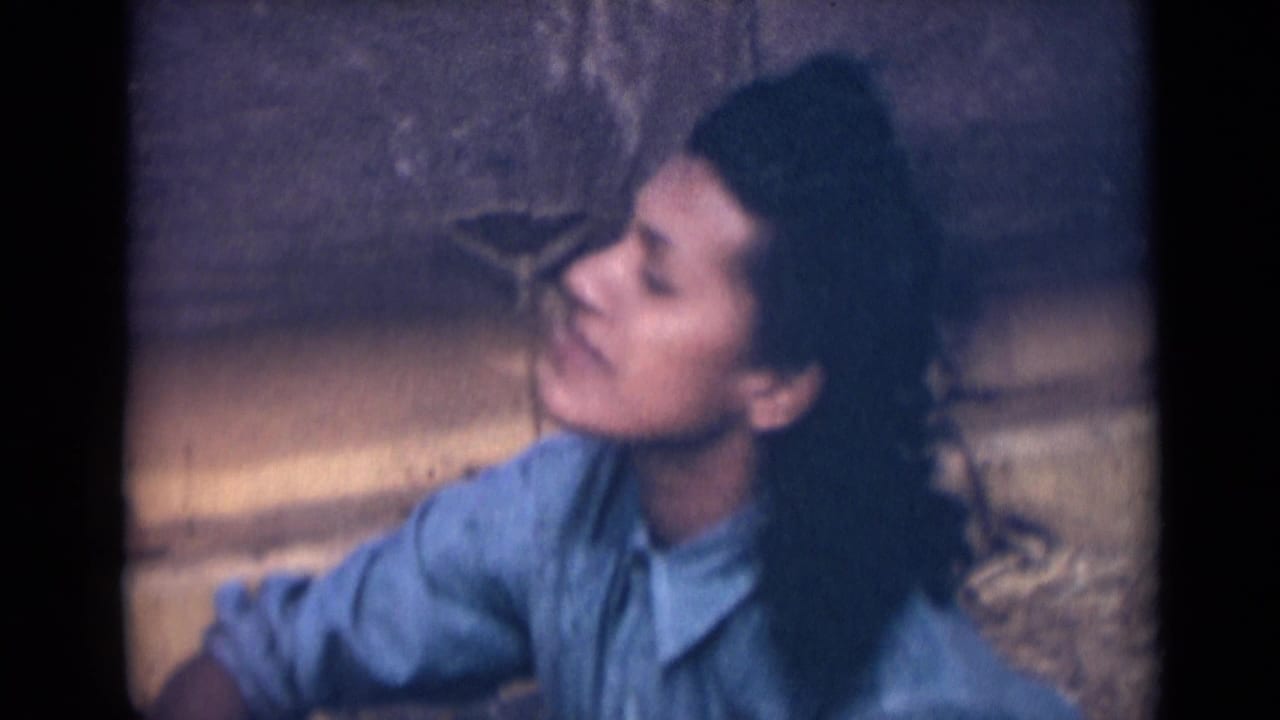 In this 1946 India 8mm frame, a young woman in a light blue collared shirt looks off camera as wind moves her dark hair near a textured stone wall.