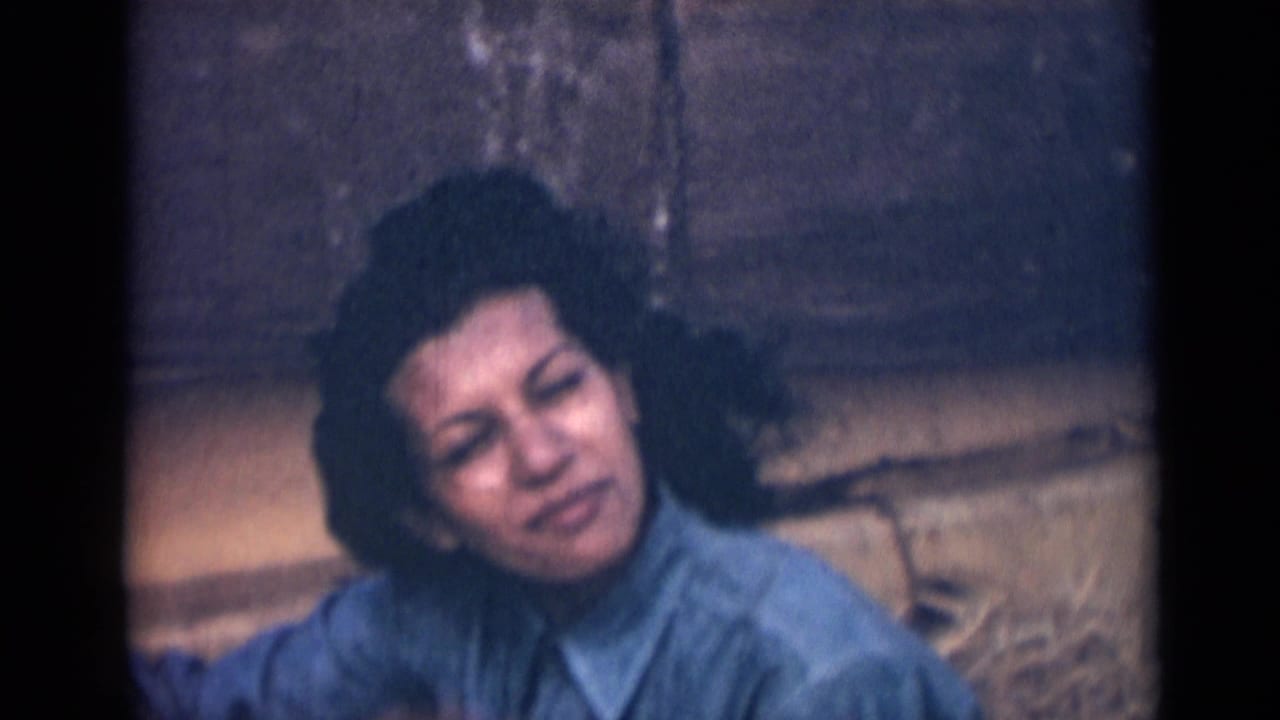 In this 1946 India 8mm frame, the woman faces closer to camera with windblown dark hair, a blue shirt, and a restrained smile against a stone wall.