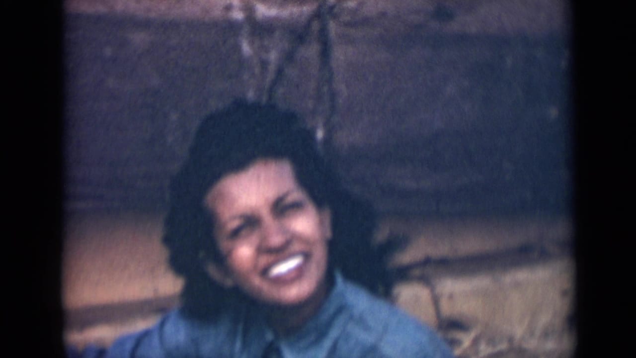 In this 1946 India 8mm frame, the woman smiles broadly toward the camera, showing teeth as her dark hair moves in the wind against the stone backdrop.
