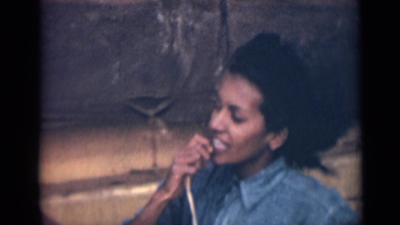 In this 1946 India 8mm frame, the woman turns in profile with a smile and a hand near her mouth, wearing a blue collared shirt beside a textured stone wall.