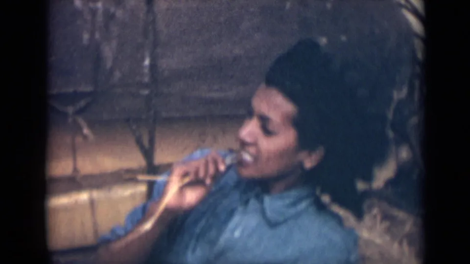 In 1946 India home-movie footage, a young woman in a blue collared shirt faces partly left while holding something near her mouth.