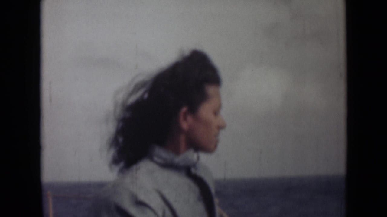 The woman remains in profile on the boat, her dark hair swept back by wind against a muted water horizon.