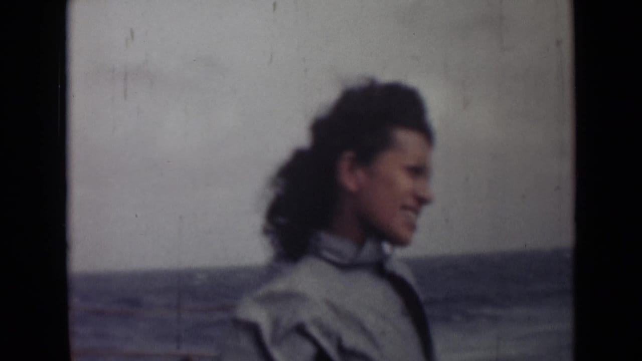 A smiling woman in a light jacket faces right as wind moves her hair, with open water visible behind her in the home-movie frame.