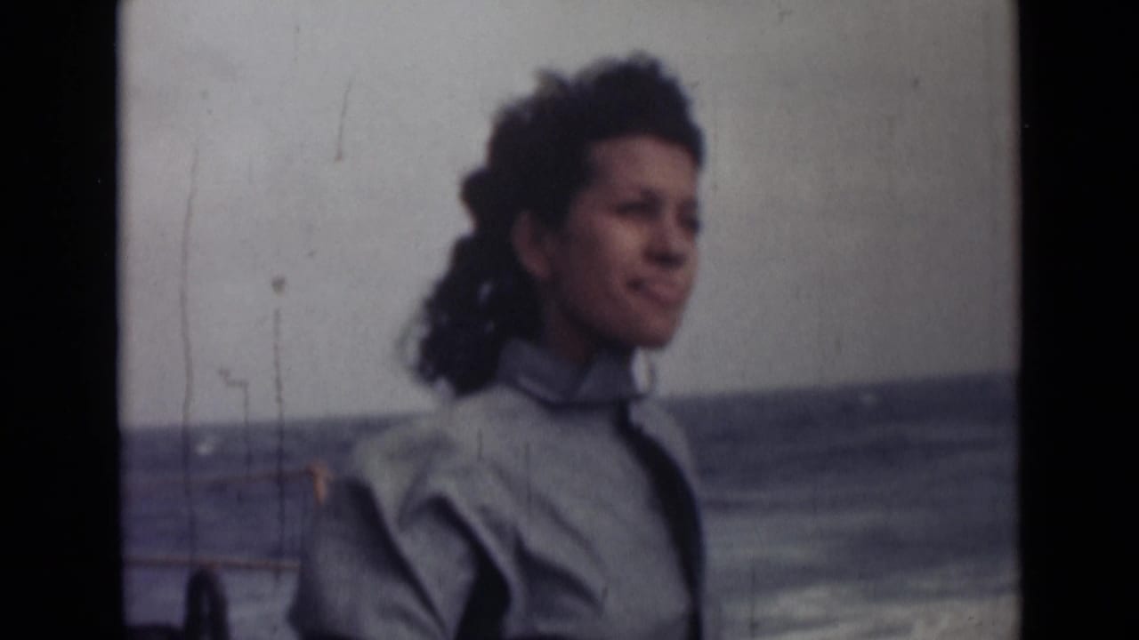 In 1946 India home-movie footage, a woman with windblown curly hair stands in profile near the ocean under a pale sky.
