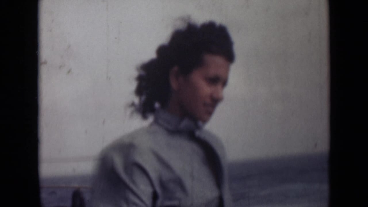 A woman in a light collared shirt appears in profile near the sea, with wind moving her dark curly hair.