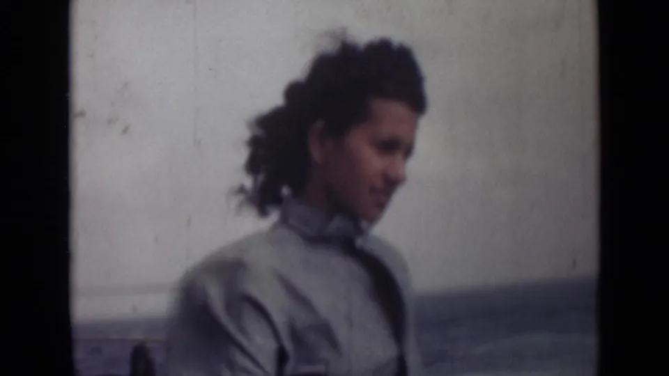 A woman in a light collared shirt appears in profile near the sea, with wind moving her dark curly hair.