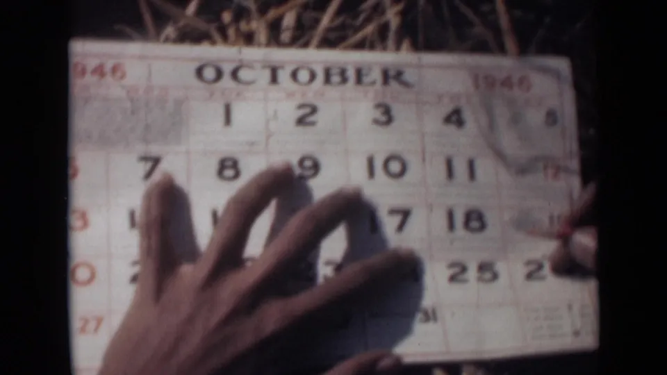 In this 1946 India 8mm frame, a hand steadies an October calendar page on straw while the date grid fills most of the view.