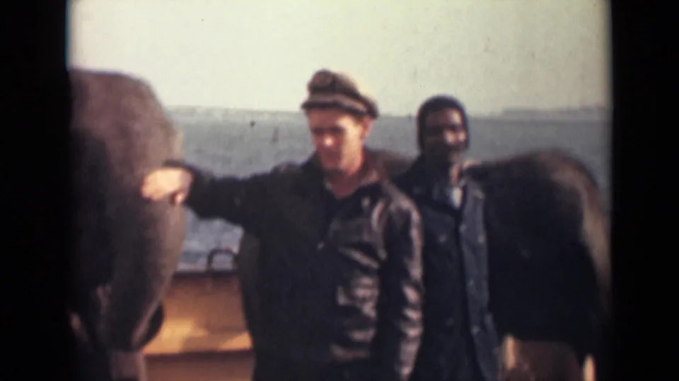 The elephant remains calm beside the capped man on a deck, with another man and open water visible in the background.