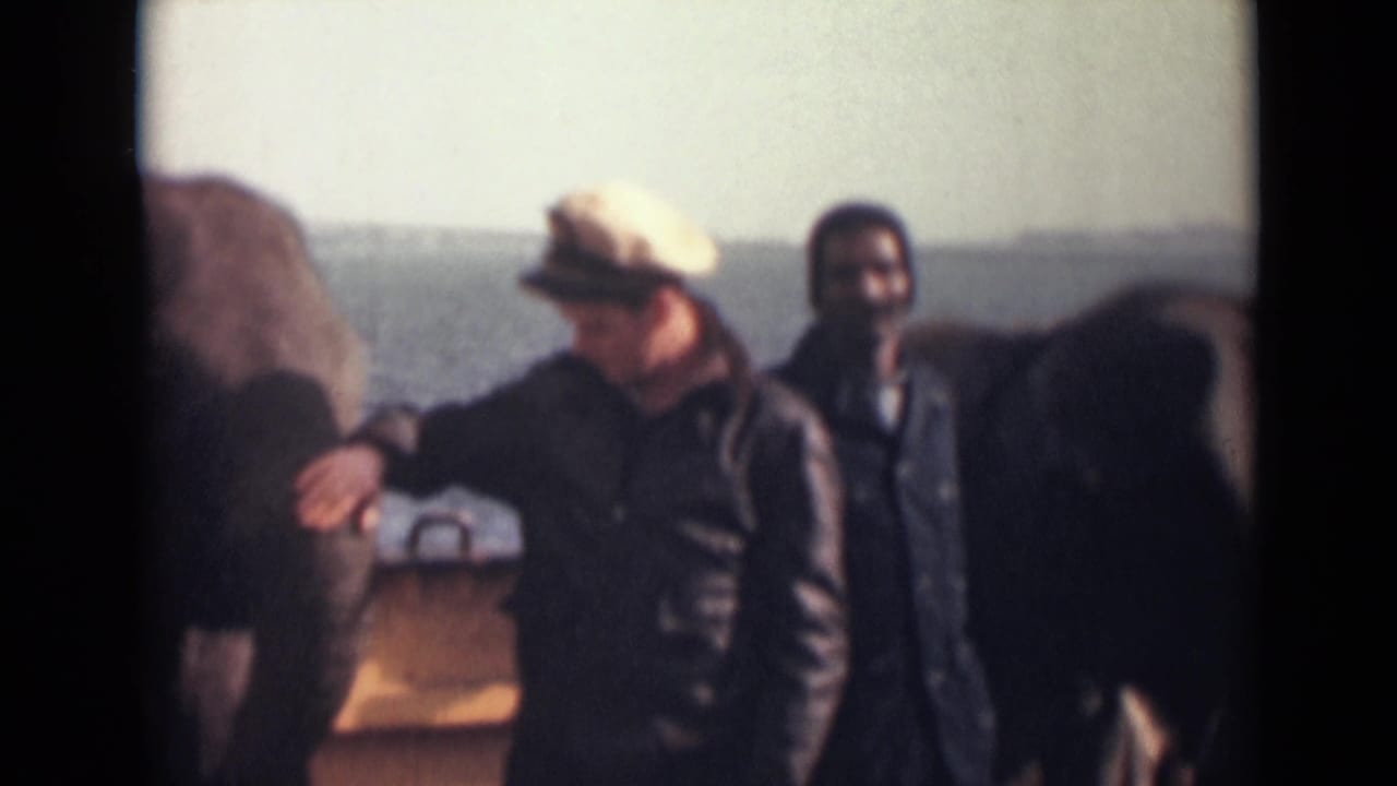 A man in a light cap turns near the elephant, with another man visible behind him and water beyond the deck rail.