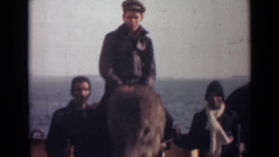 1946 India home-movie frame with an elephant close to camera, a capped rider, and two men posed against the sea.