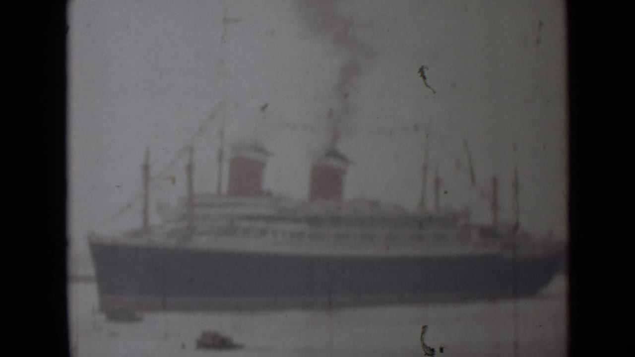 Passenger ship with two funnels and smoke, viewed across calm harbor water.