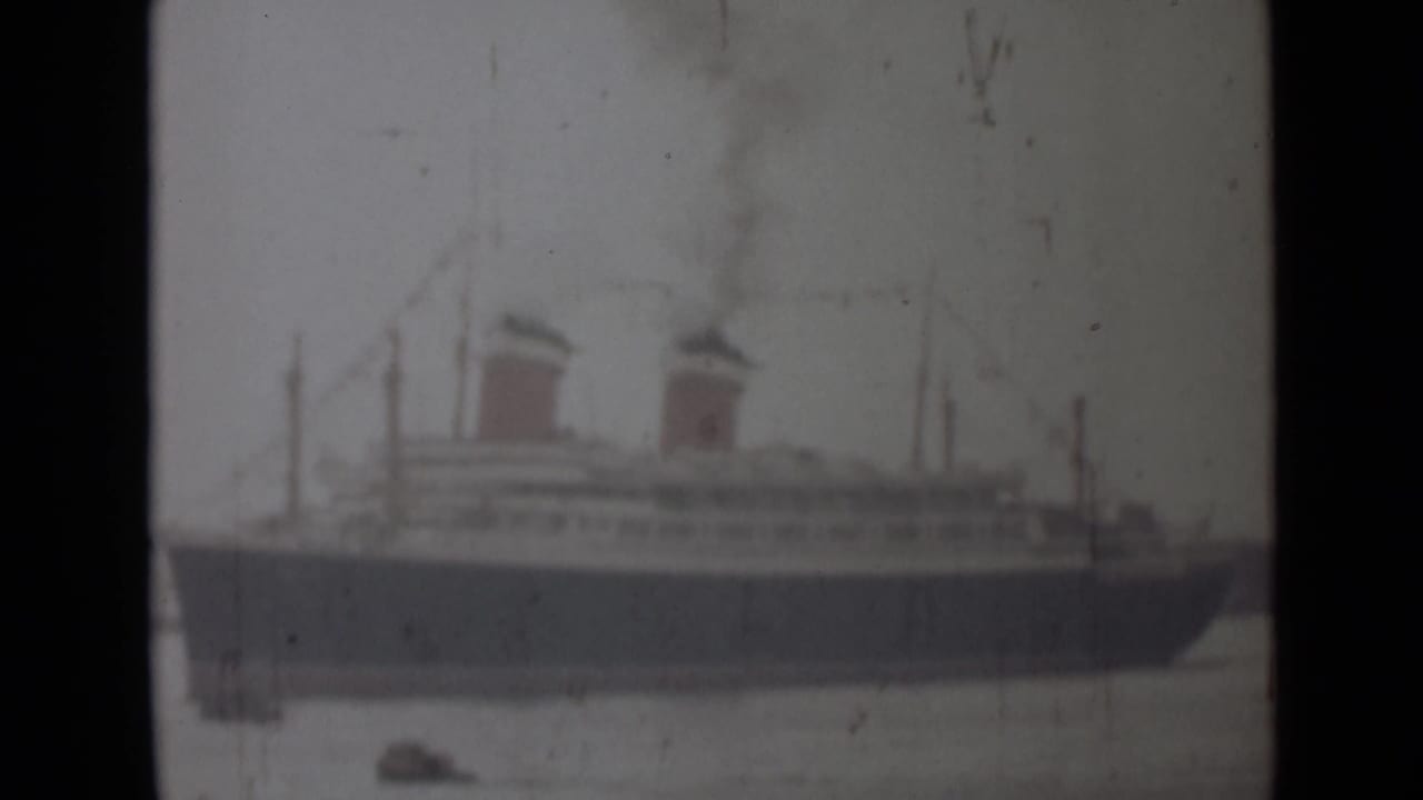 Faded 1946 India home-movie frame showing the two-funneled passenger ship closer in view, with smoke and small boats below.