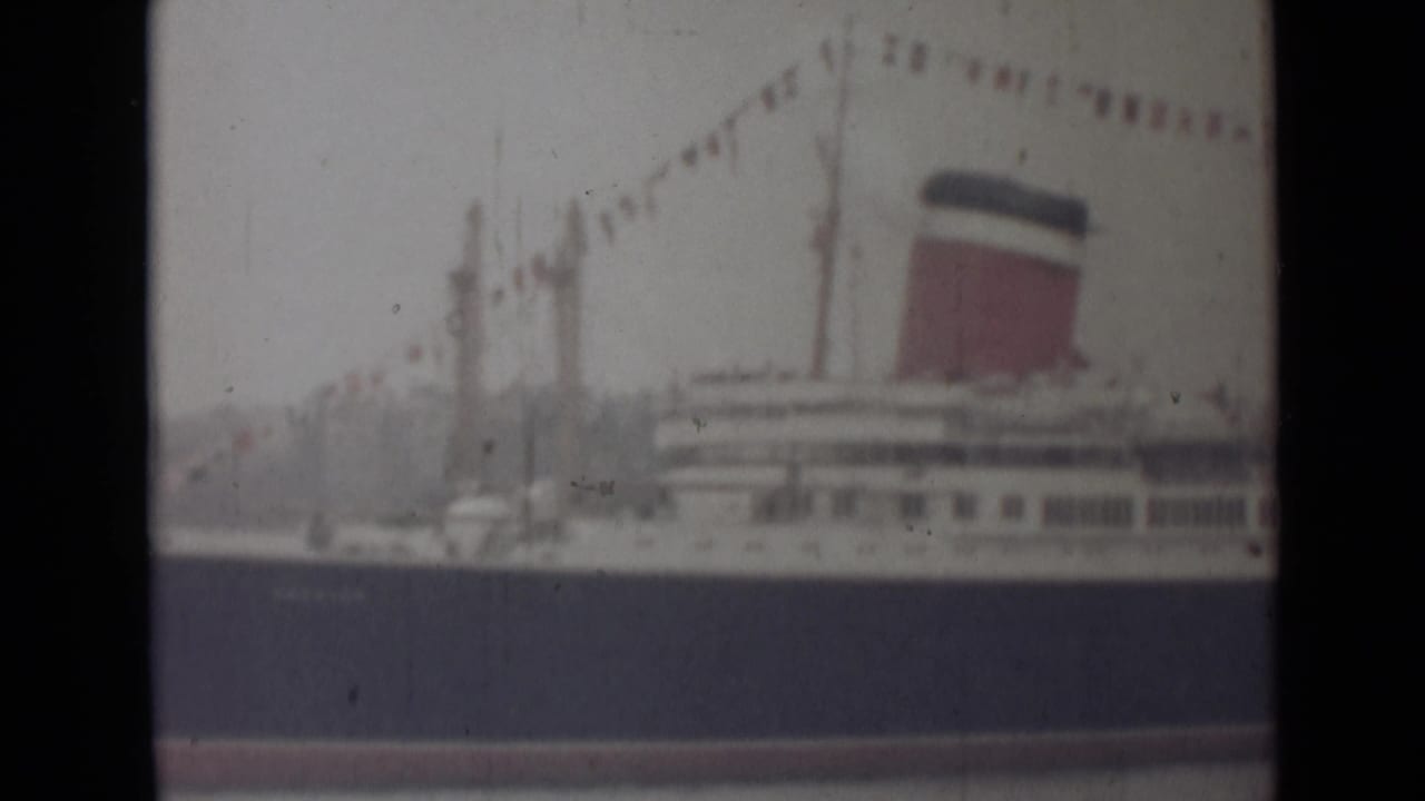 1946 India 8mm frame showing a close view of the liner's funnel, upper decks, and bunting against a hazy harbor backdrop.
