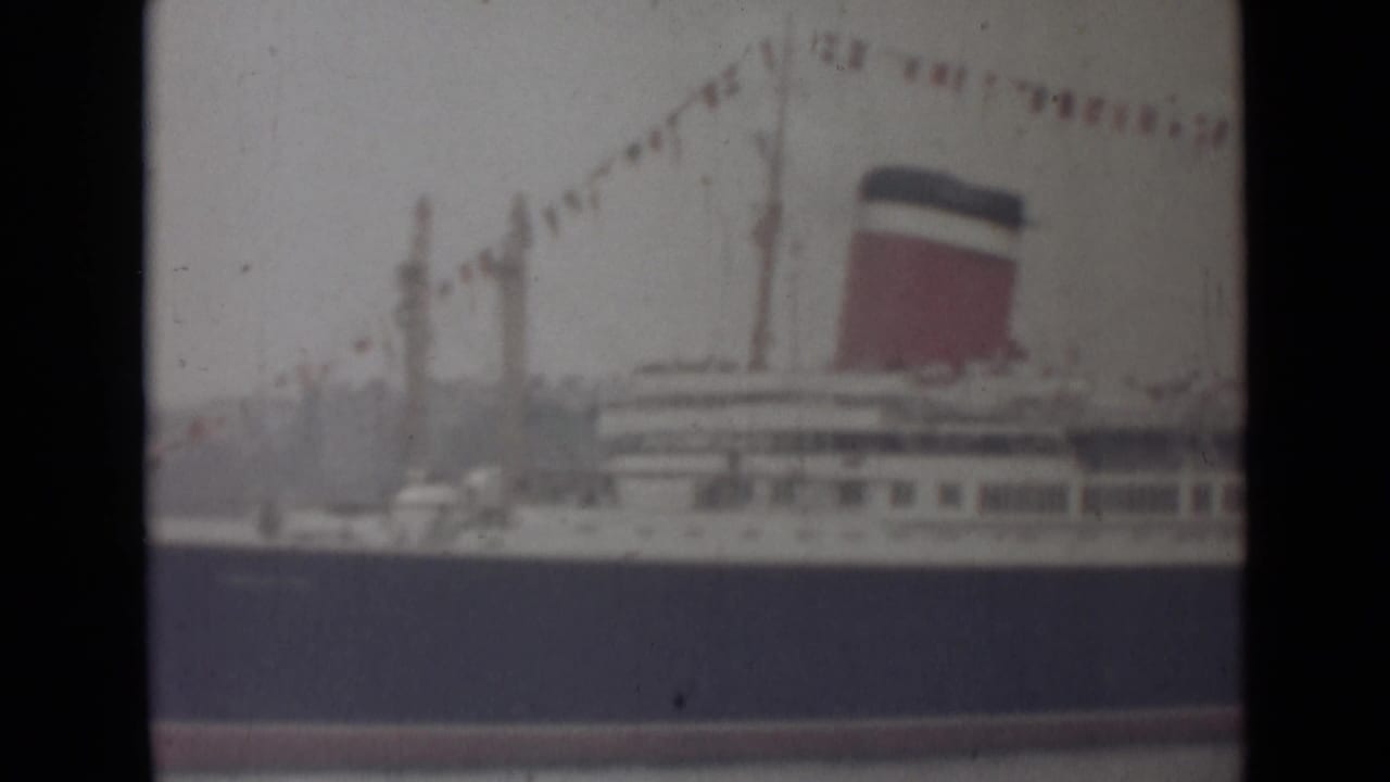 1940s India home-movie frame of a large white ocean liner with a red-and-dark funnel and signal flags above calm water.