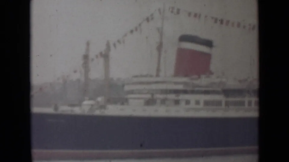 1940s India home-movie frame of a large white ocean liner with a red-and-dark funnel and signal flags above calm water.