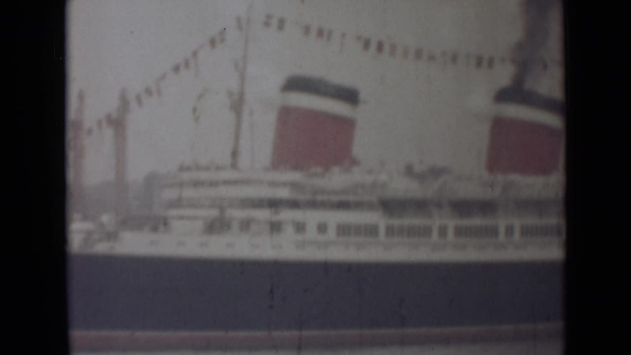 Faded 8mm frame of the India-location liner with flags strung above, deck windows below and two red-and-dark funnels visible.
