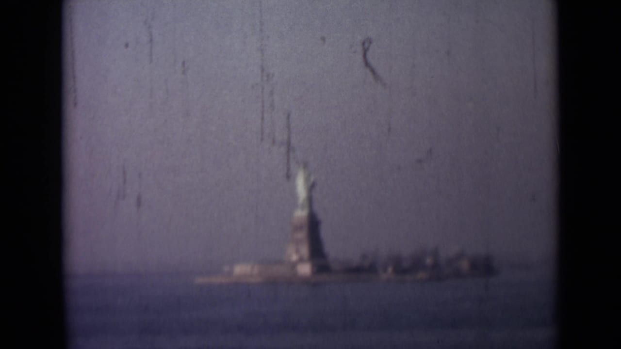 Distant 8mm harbor view of the Statue of Liberty rising above the water, filmed from a moving vessel.