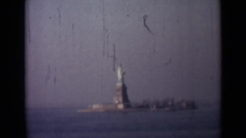 Distant 8mm harbor view of the Statue of Liberty rising above the water, filmed from a moving vessel.
