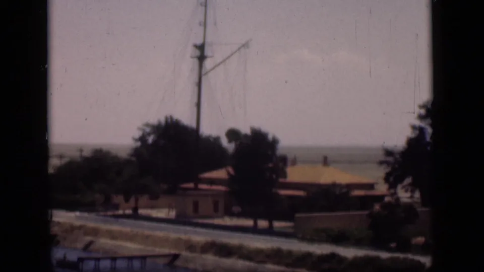 1946 Suez Canal, Egypt home-movie frame showing a quiet canal-side area with palms, low buildings, a road, and a tall mast.