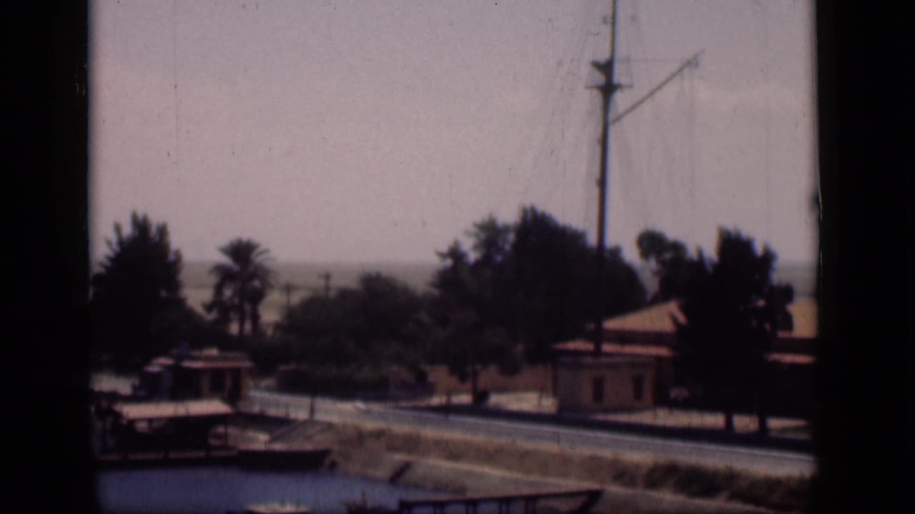1946 Suez Canal, Egypt home-movie frame showing water in the foreground, roadside buildings, palms, and a tall mast.