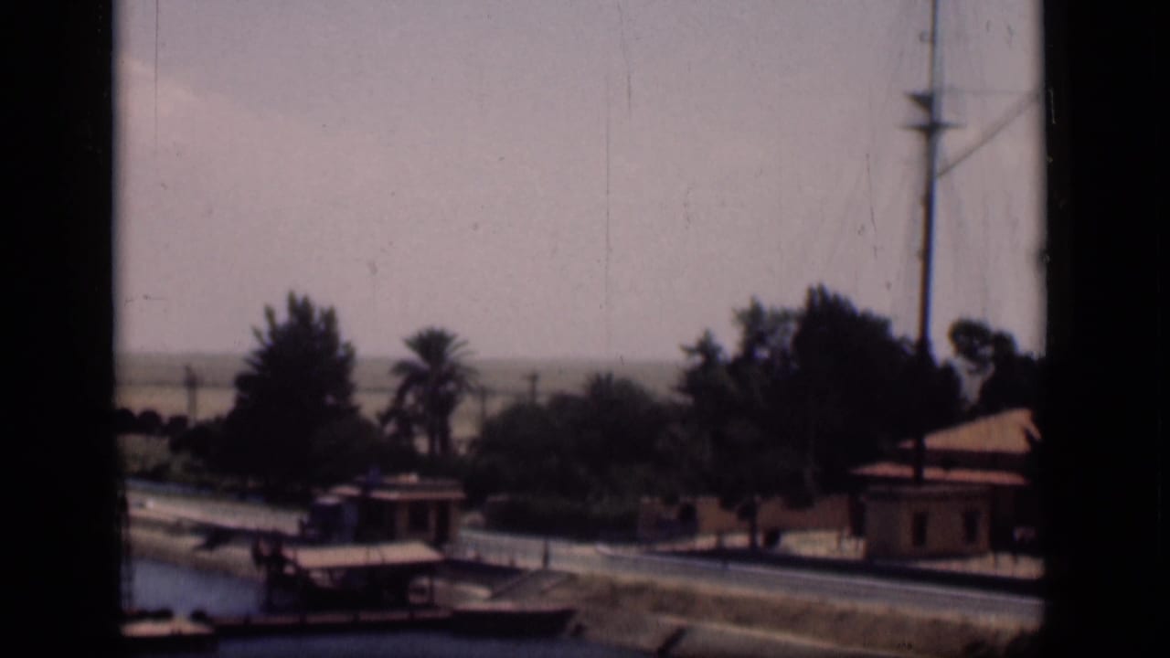 1946 Suez Canal, Egypt frame showing low canal-side structures, palms, a road, and a mast near the right edge.