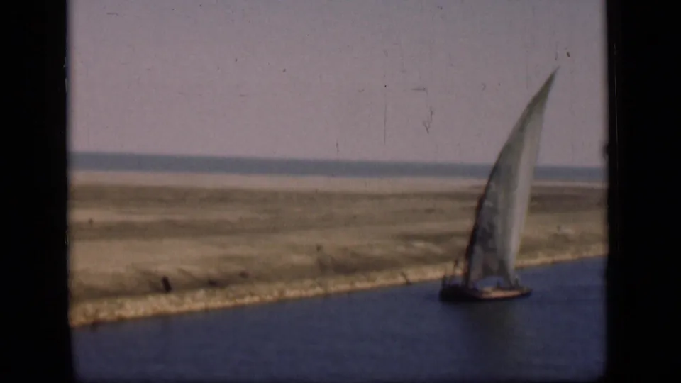 A sailboat with a tall white sail moves along the Suez Canal area in 1946, with grassy banks and open land beyond.