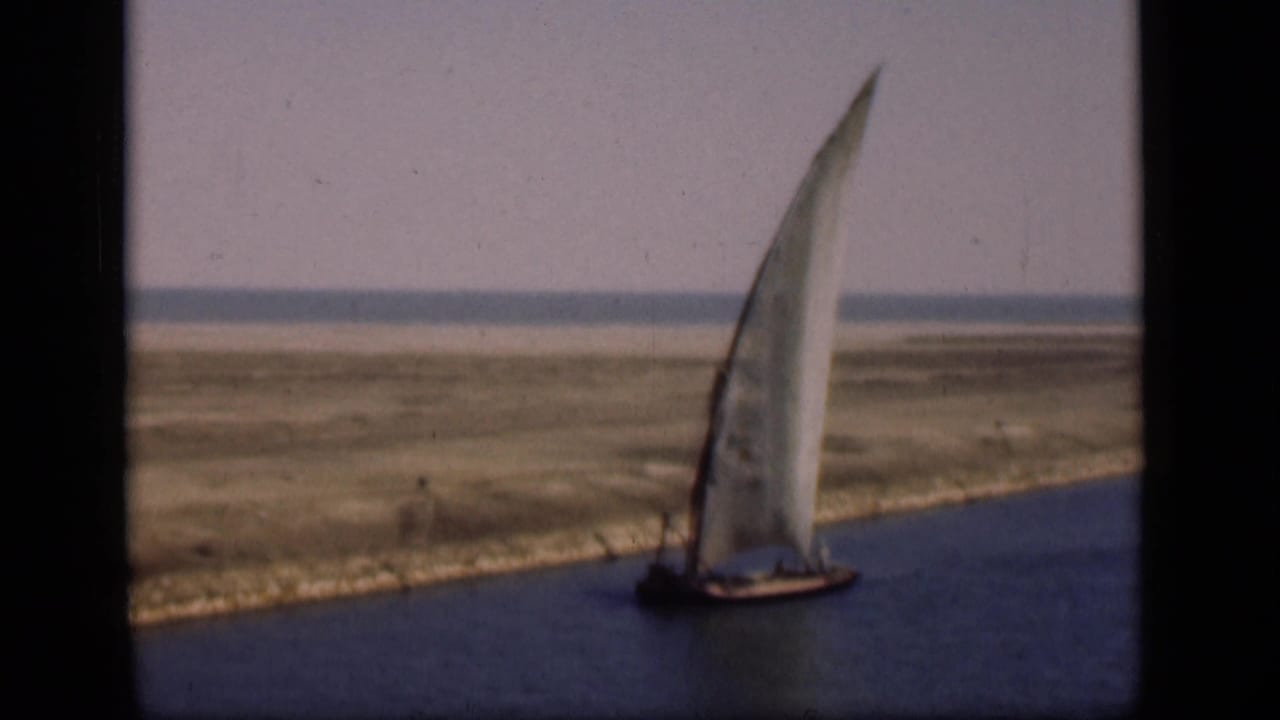 1946 Suez Canal home-movie frame showing a tall-sailed boat on blue water with a broad sandy plain and pale sky behind it.
