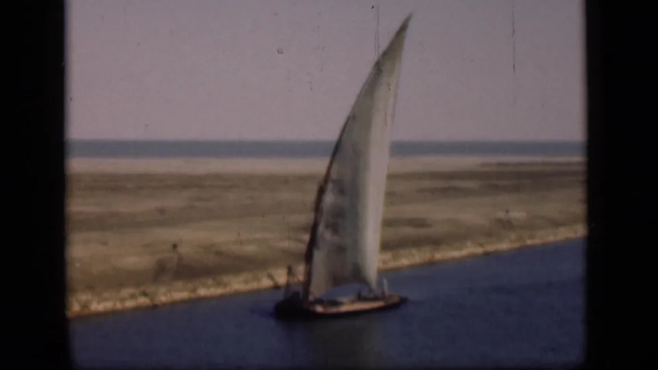 1946 Suez Canal home-movie frame showing a felucca with a large lateen sail on blue water near flat canal banks.