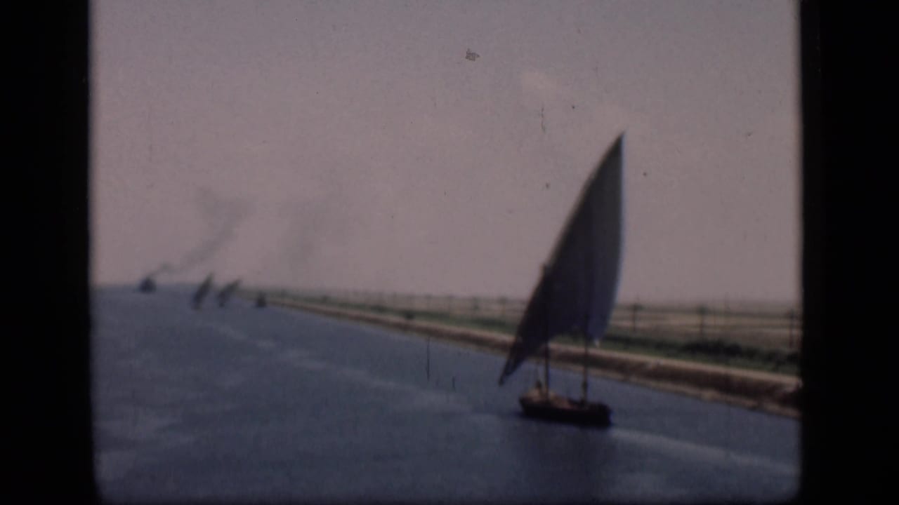 Lateen sailboat on blue canal water with low banks and distant smoke.