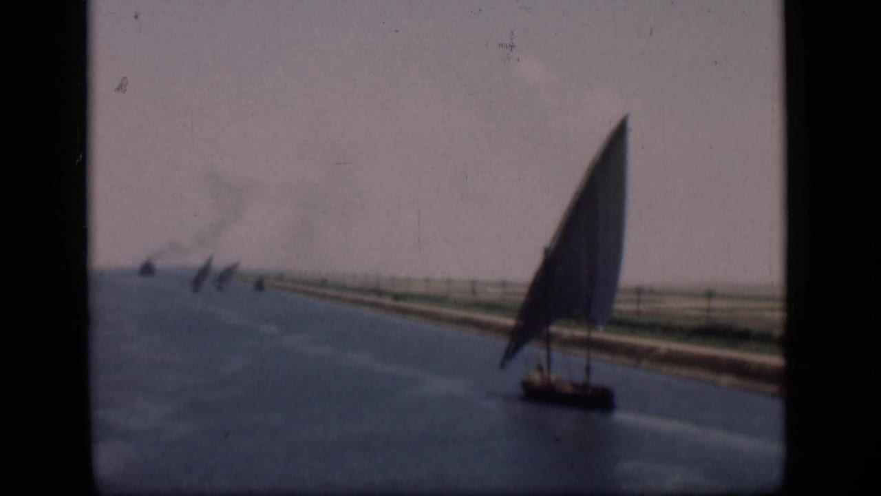 Soft 1946 Suez Canal home-movie frame with a lateen sailboat, blue water, low shoreline, distant sail traffic, and visible film grain.