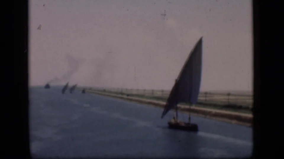 Soft 1946 Suez Canal home-movie frame with a lateen sailboat, blue water, low shoreline, distant sail traffic, and visible film grain.