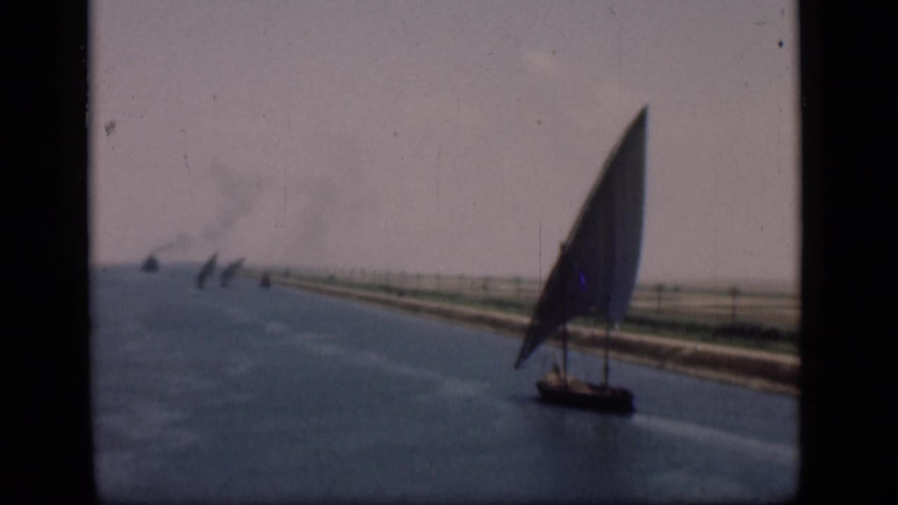1946 Suez Canal frame showing a lateen sailboat on blue water, flat canal banks, distant sails, faint smoke, and aged 8mm color.