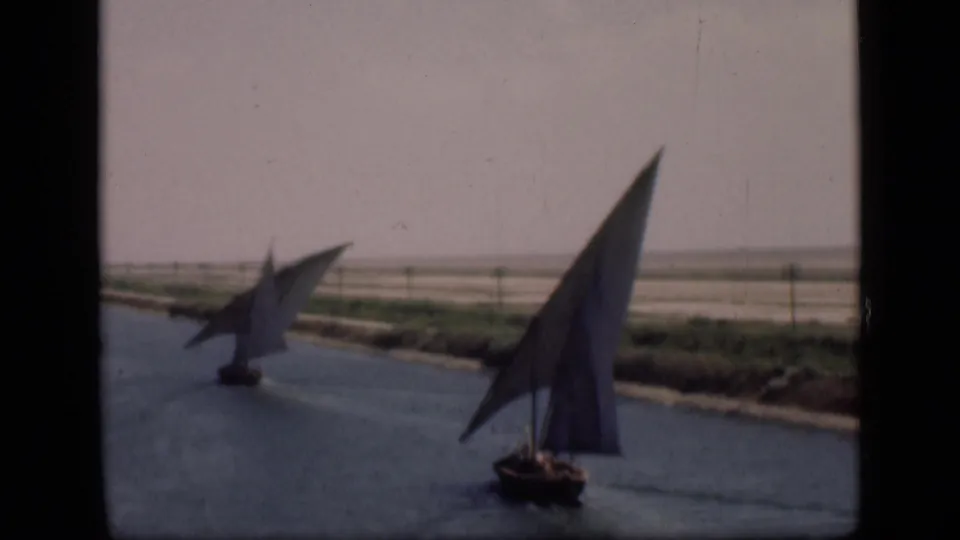 Two felucca sailboats appear on the Suez Canal, one close to camera and another crossing farther back.