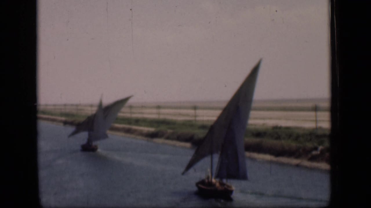 1946 Suez Canal footage frame with two sailboats under triangular sails moving past a flat, arid shoreline.