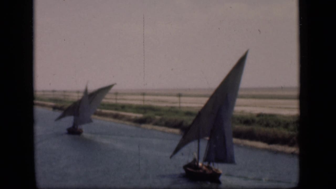 Two sailboats with triangular sails move along the Suez Canal in Egypt, with flat banks and hazy sky in the background.