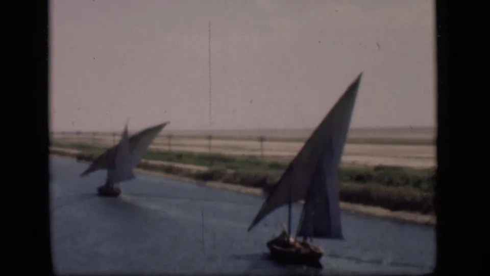 Two sailboats with triangular sails move along the Suez Canal in Egypt, with flat banks and hazy sky in the background.