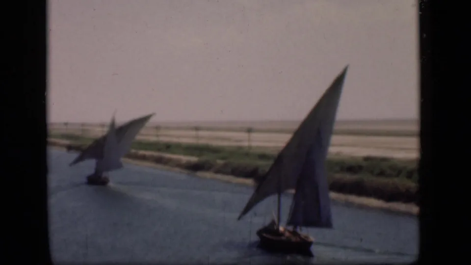 A pair of felucca-style boats crosses the Suez Canal, their sails silhouetted against pale banks and open sky.