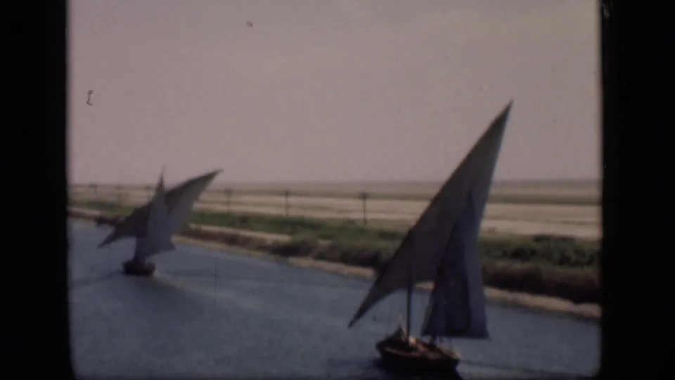 Two traditional sailboats are seen from an elevated or bank-side view on the Suez Canal, with open land beyond the water.
