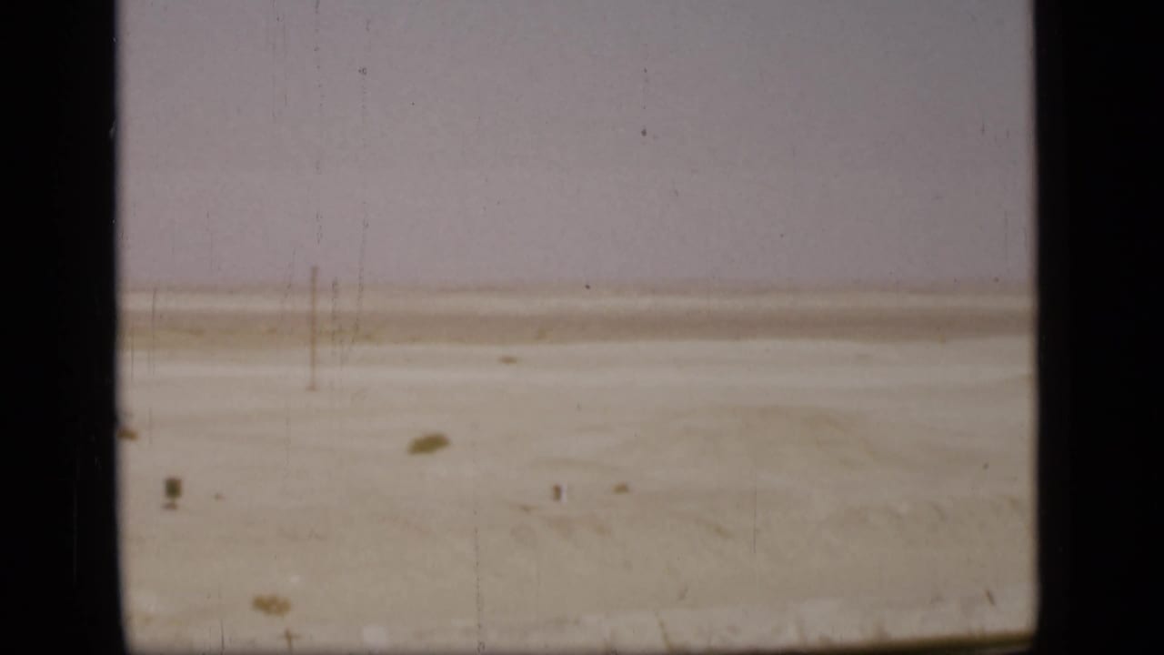 Flat pale desert with sparse scrub under a washed sky, seen through 8mm gate edges.