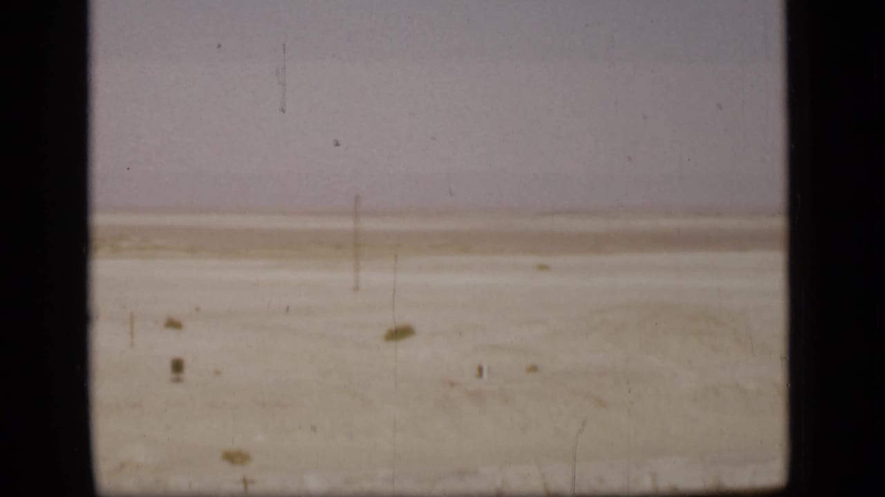 Wide sandy flat with a faint horizon, small scrub, and vertical film scratches.