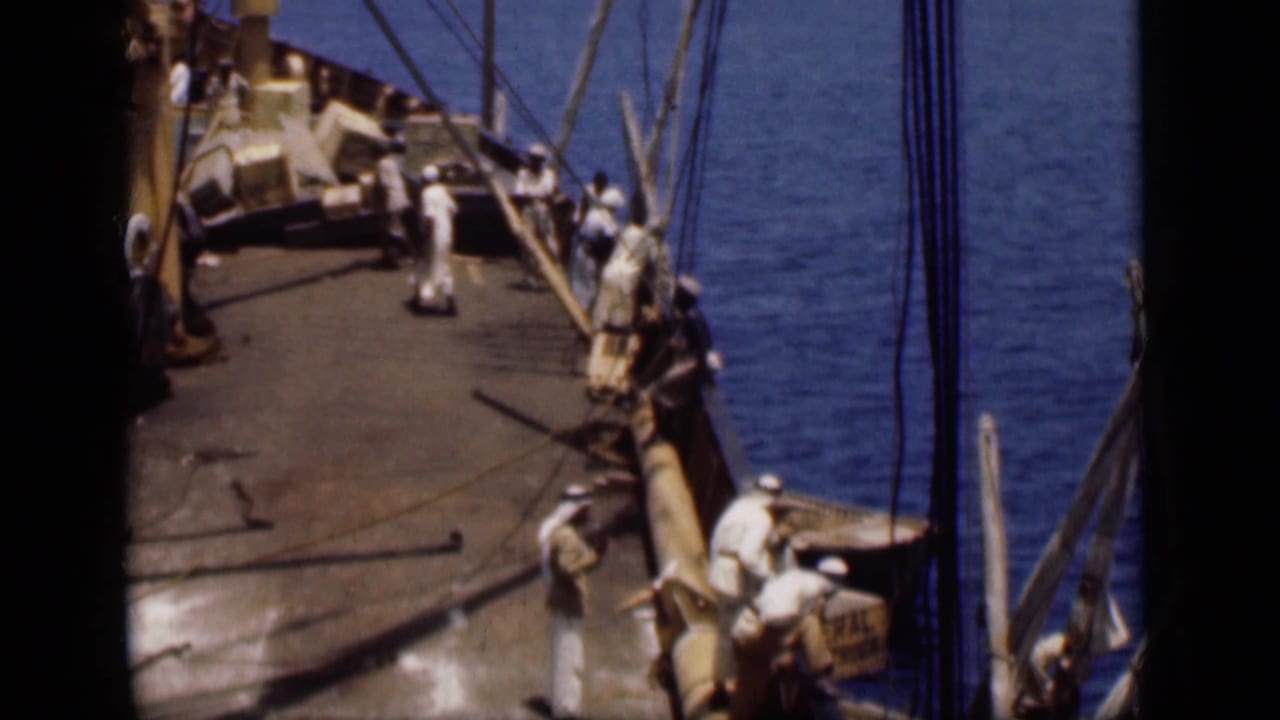 Men in white clothing move on a ship deck along the Suez Canal, with rigging and blue water visible beside them.