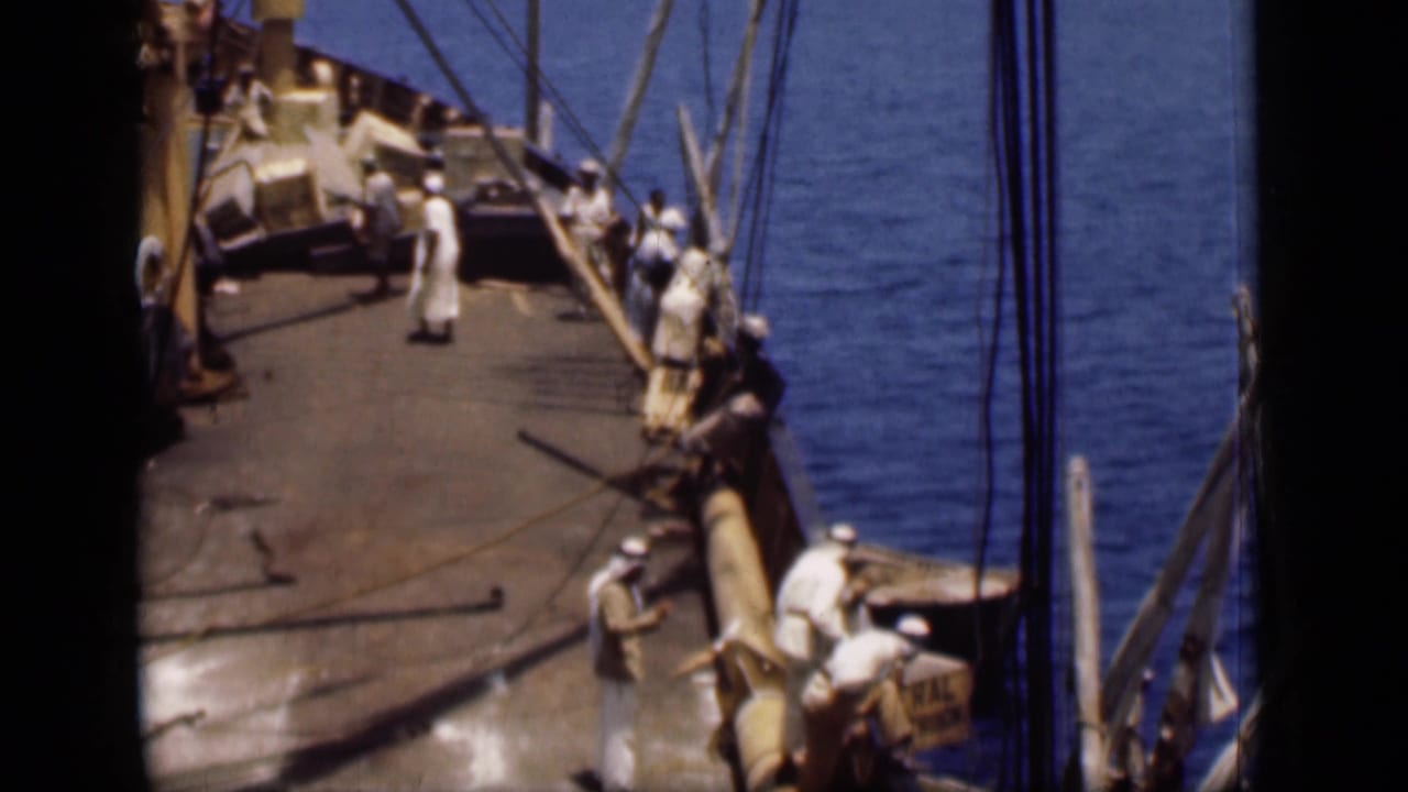 Crew members on a wooden sailing vessel deck with rigging and blue water alongside.