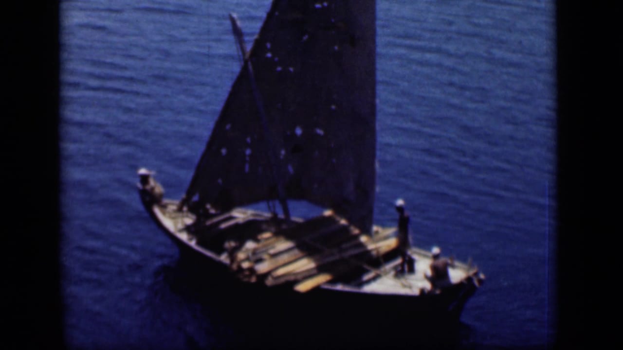 Small wooden sailboat with a dark triangular sail crosses blue water.