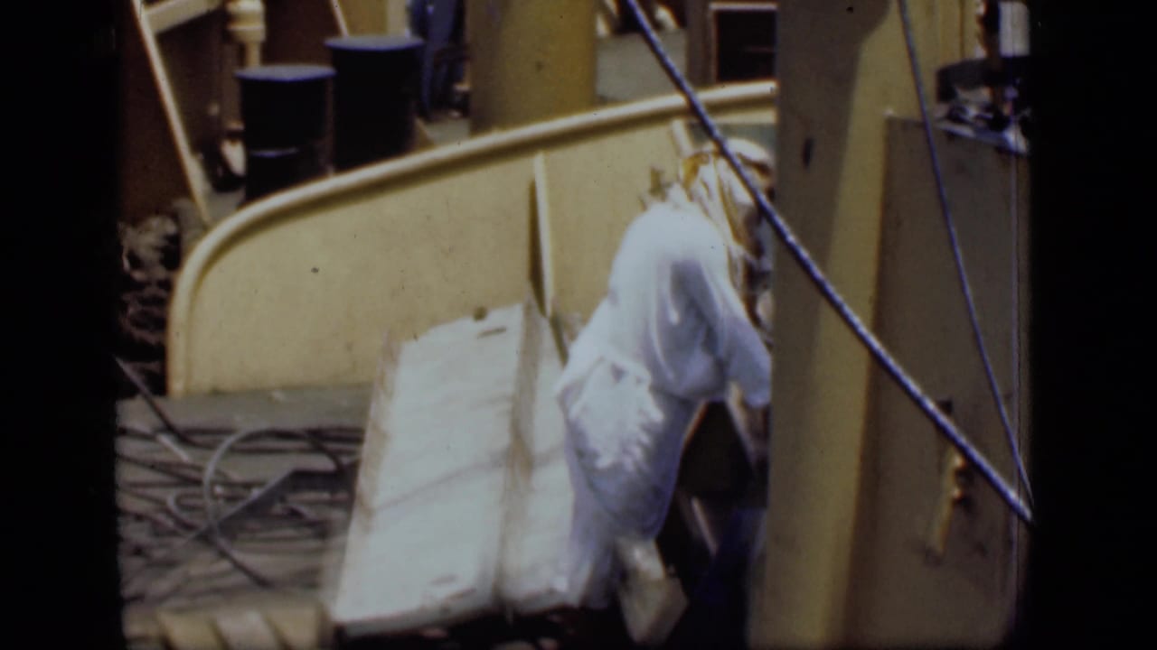 1946 Suez Canal home-movie frame showing a man in white clothing leaning over boards or scrap on a sunlit dock area.